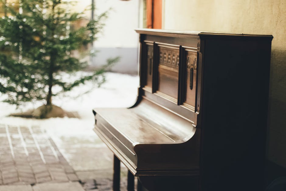Close-up of a vintage upright piano with a dark wooden finish, showing the white and black keys extending across the keyboard. The piano has a brass hinge along the top edge where the lid opens, and a