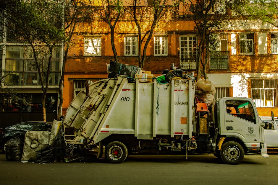 Two green outdoor waste bins with black lids are positioned side by side on a concrete pavement at the edge of a street, adjacent to a small pile of debris and some paving slabs. Behind the bins, a se